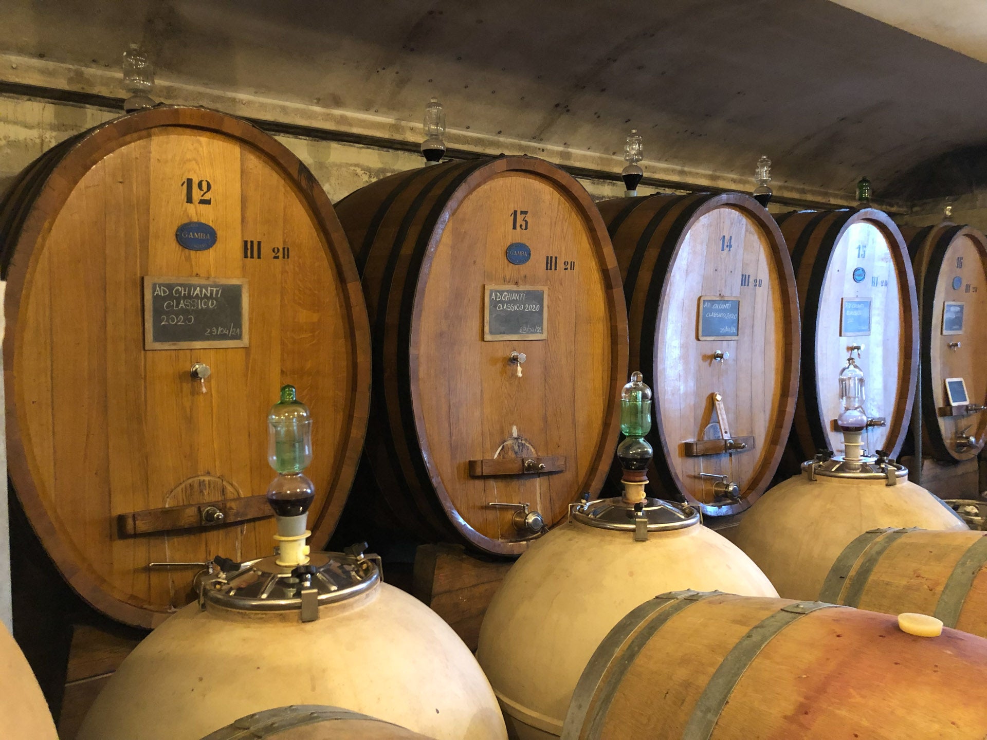 Large wooden wine barrels labeled with numbers 12, 13, and 14 are lined up in a cellar at the best wine shop in Nashville. Smaller barrels are in front of them. Labels on the large barrels indicate "Chianti Classico 2022." Sealed airlocks are attached to the barrels for fermentation.