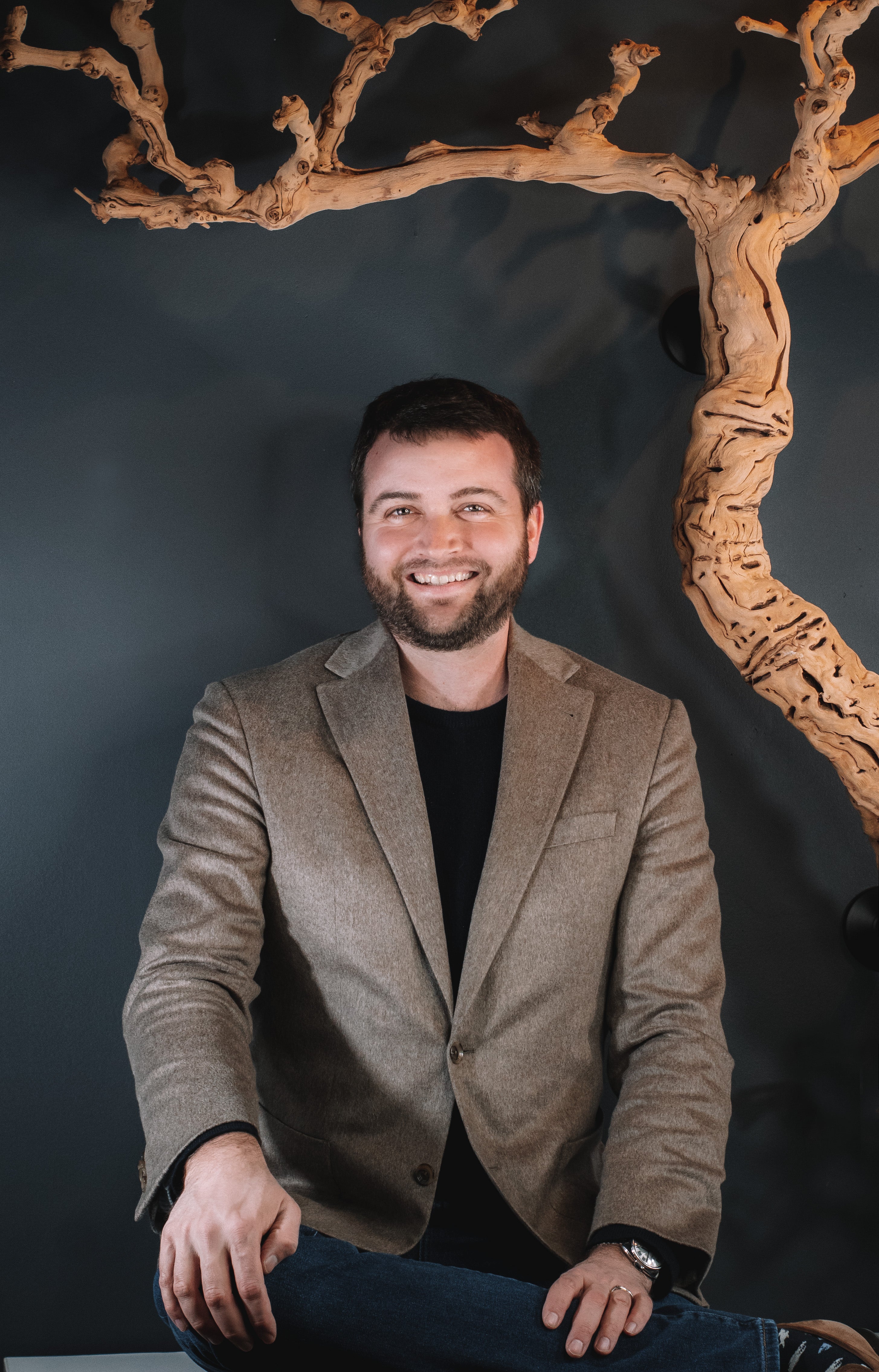 Brian,  man with short dark hair and a beard smiles while seated. He is wearing a brown blazer over a black shirt and blue jeans,  in front of a dark blue wall adorned with a twisted grape vine in Nashville's Wine and Spirits store, Harvest Wine Market.