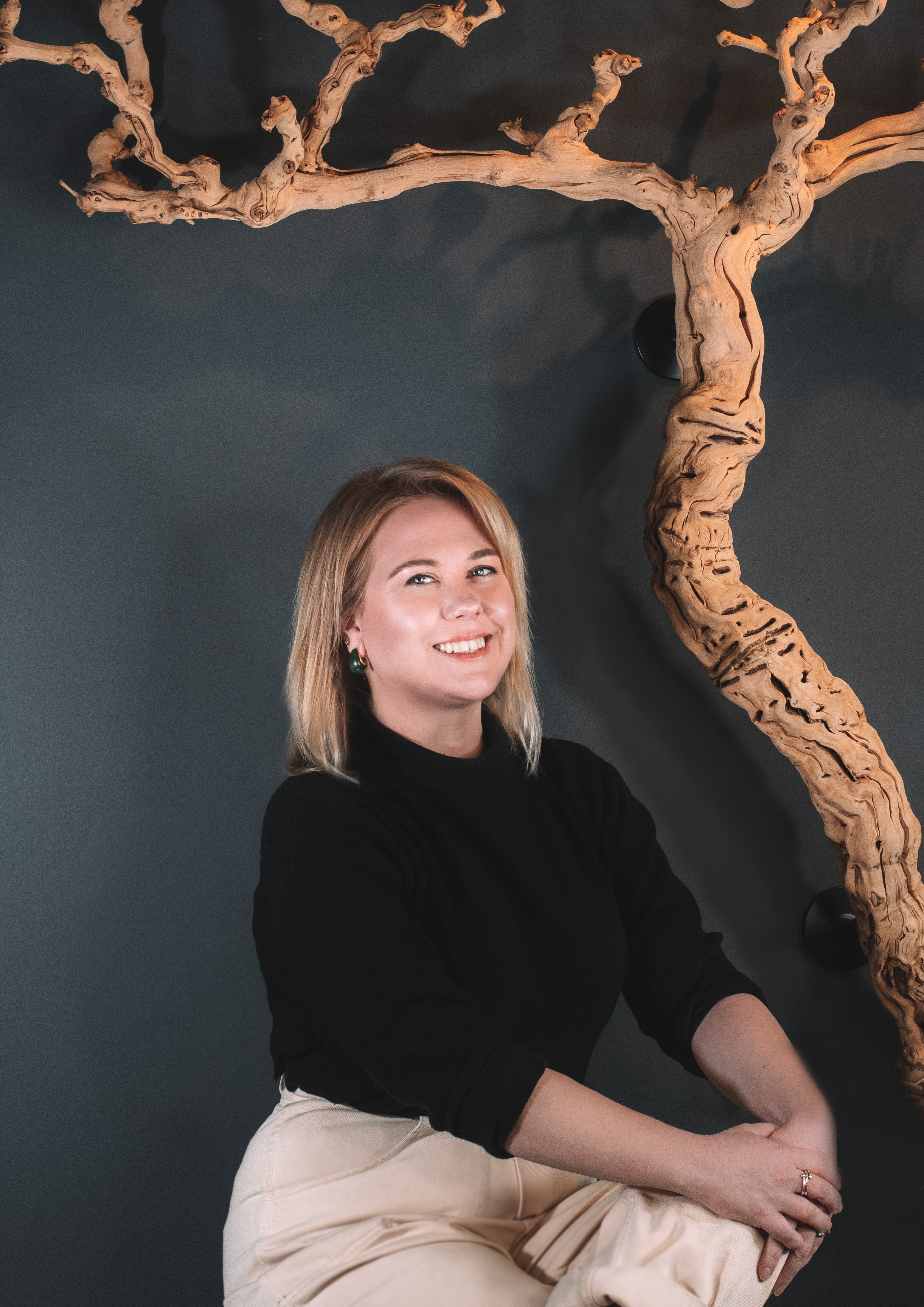 LeeAnn, a person with shoulder-length blonde hair sits in front of a dark blue wall adorned with a twisted grape vine in Nashville's Wine and Spirits store, Harvest Wine Market.