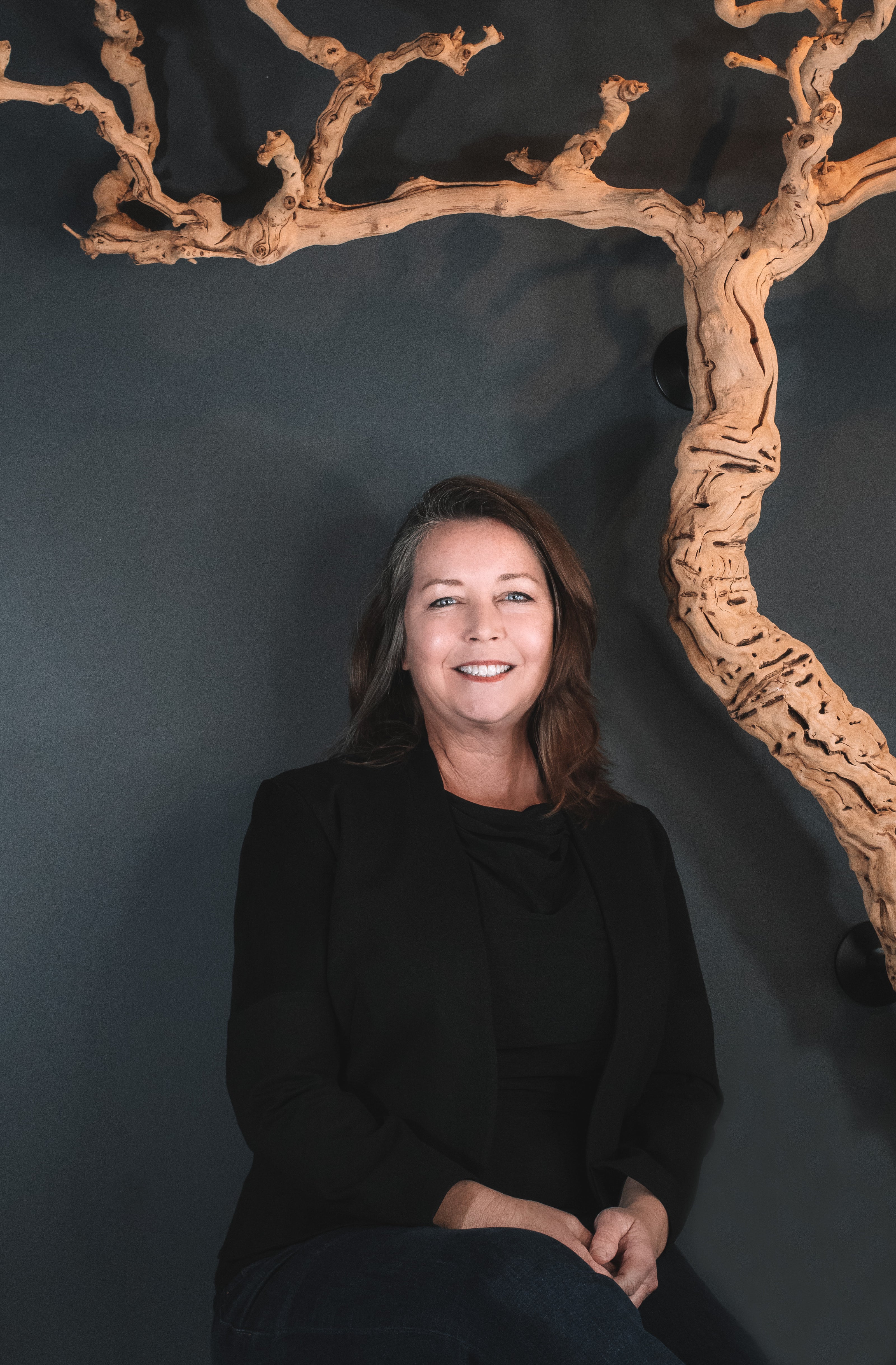 Sarah is smiling with long brown hair, wearing a black blazer and shirt, sitting in front of a dark blue wall adorned with a twisted grape vine in Nashville's Wine and Spirits store, Harvest Wine Market.