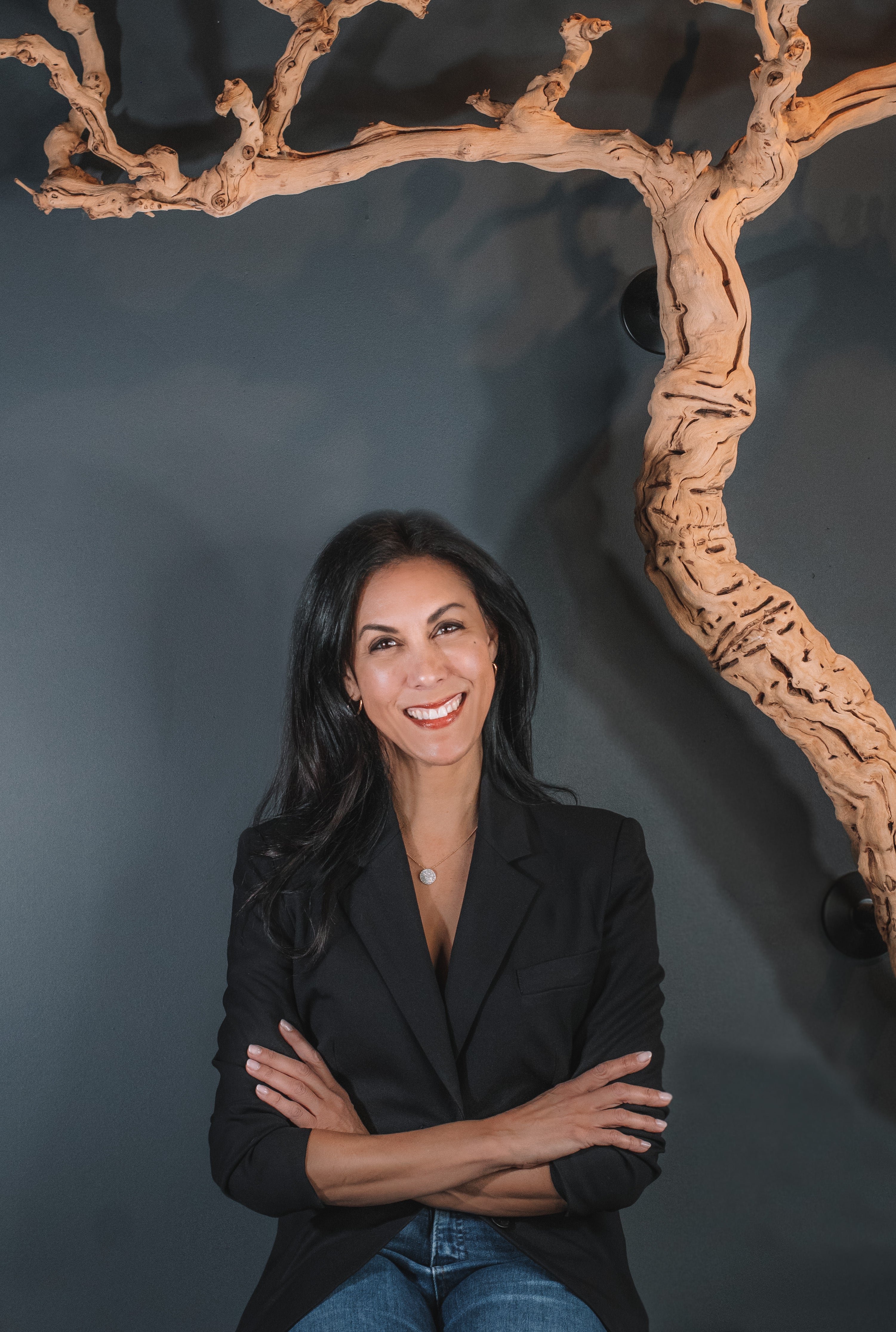 Seema, a person with long dark hair, wearing a black blazer and jeans, is smiling with their arms crossed. They are sitting in front of a dark blue wall adorned with a twisted grape vine in Nashville's Wine and Spirits store, Harvest Wine Market.