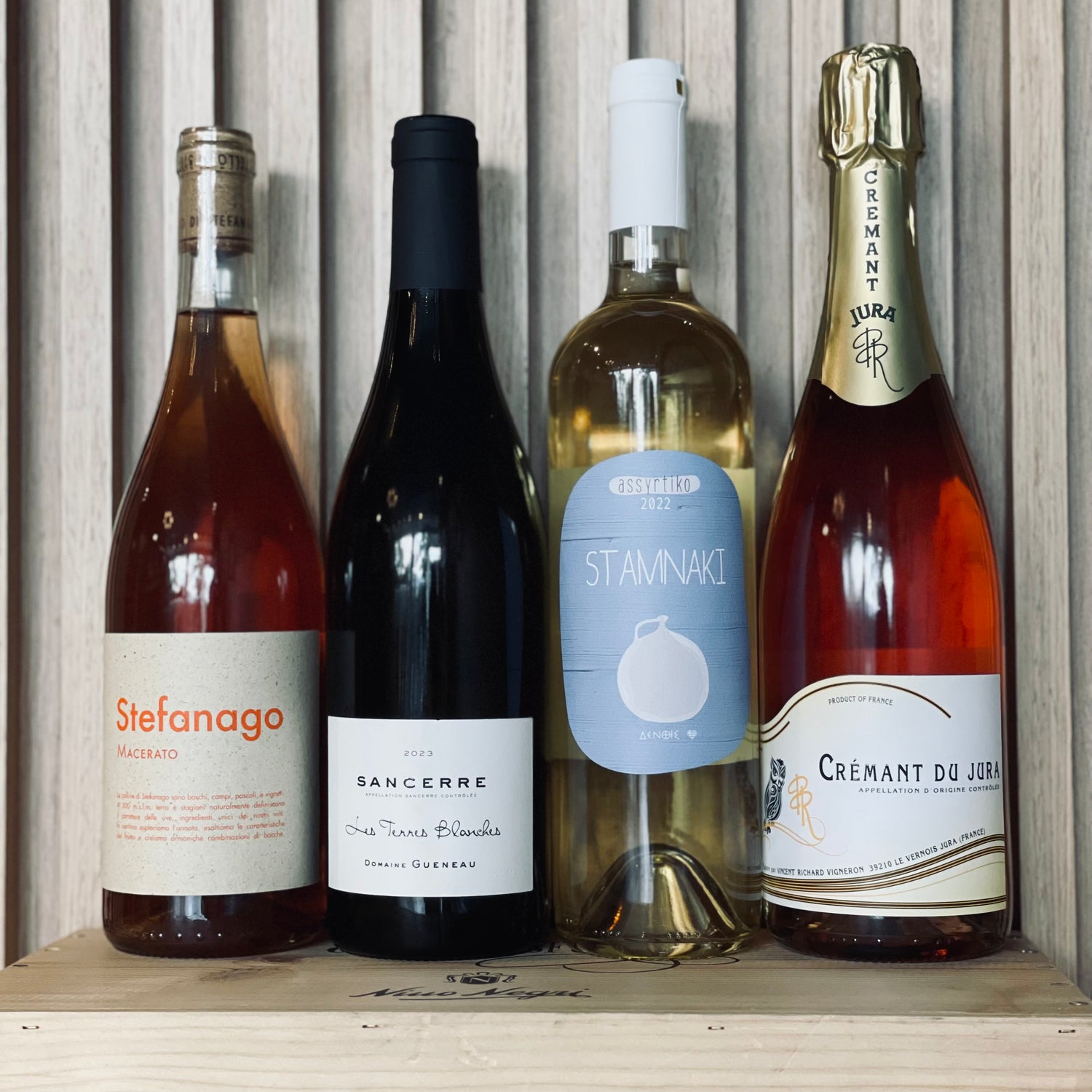 A wooden crate displaying four bottles of wine against a wooden backdrop, highlighting the offerings from Wine and Spirits Nashville TN. From left to right: a bottle of Stefanago, Sancerre, Stamnakis, and Cremant du Jura. Each bottle boasts distinctive labels and colors.